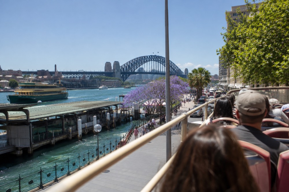 An open-top sightseeing bus on a Sydney street with the harbour bridge in view