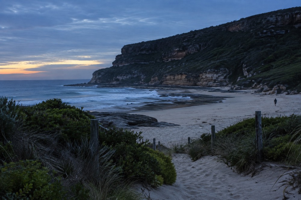A calm ocean beach at the foot of sandstone cliffs with a sandy walking track leading to the sand