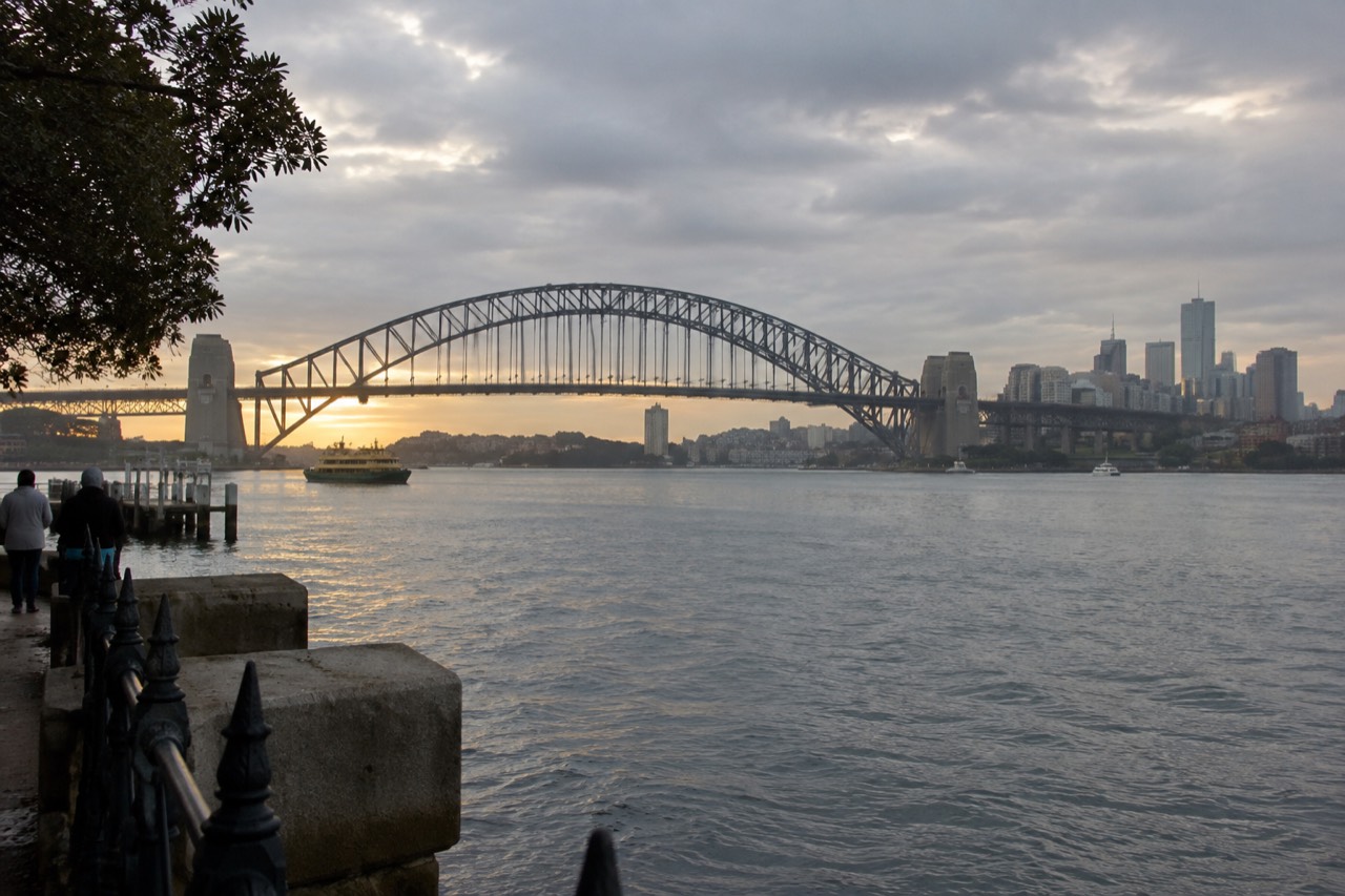 The steel arch of Sydney Harbour Bridge rising above the water at dawn with the city skyline behind it