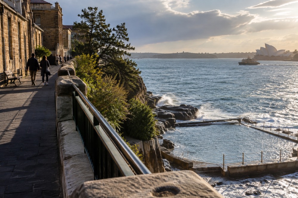 A paved clifftop path above a small ocean pool with waves breaking on the rocks below
