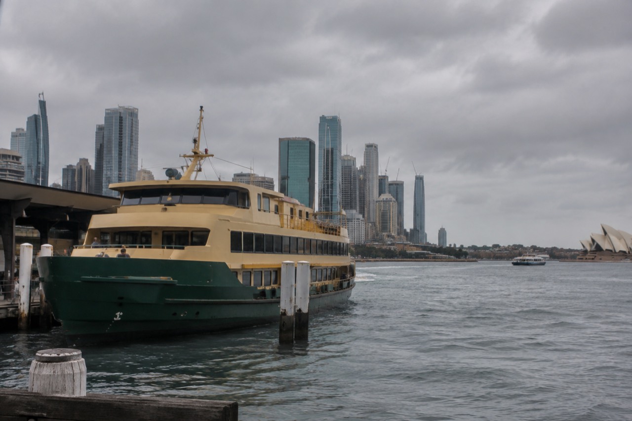 A green and yellow Sydney ferry at Circular Quay wharf with the city skyline behind it