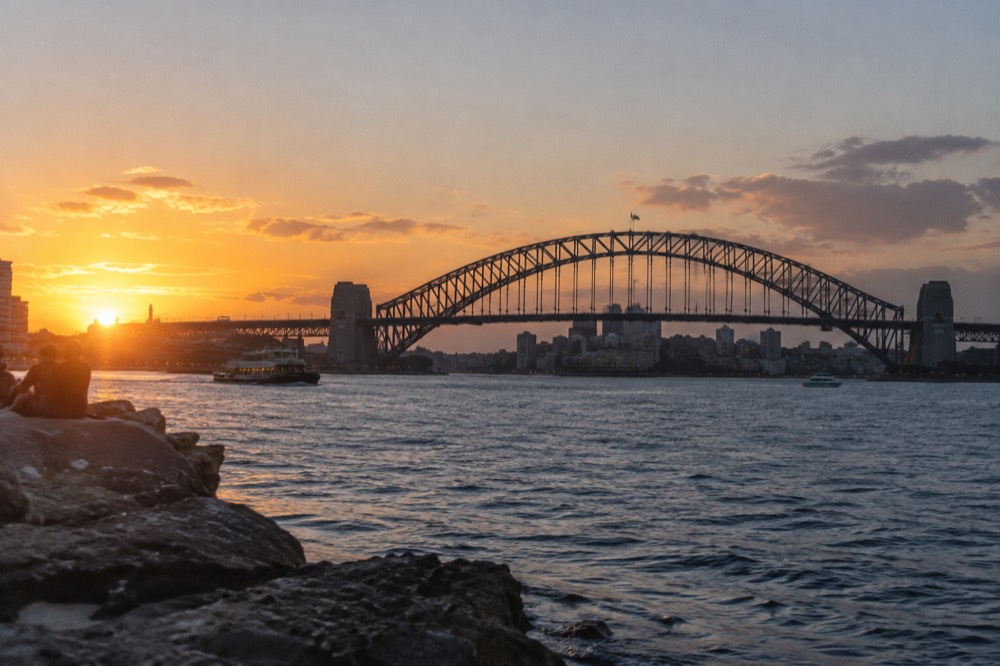 The steel arch of Sydney Harbour Bridge seen from the harbour at dusk with city lights behind