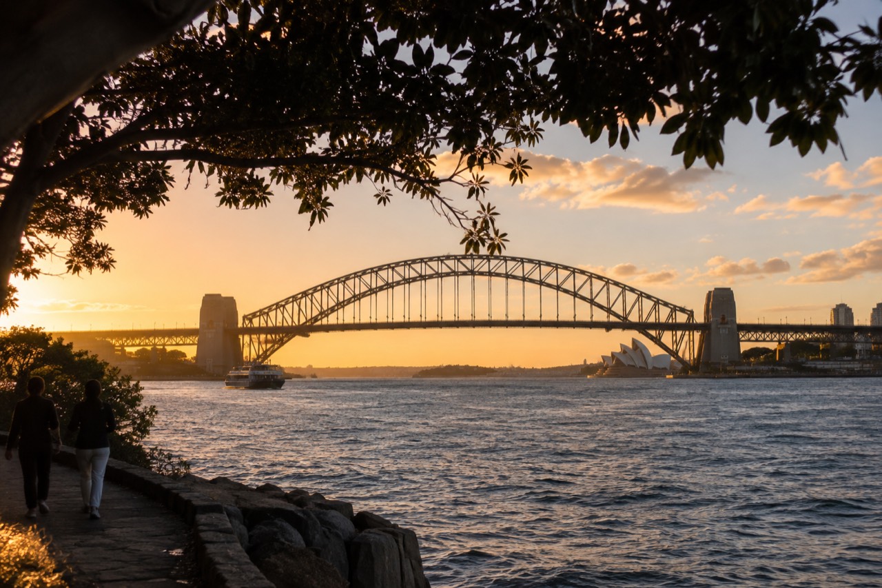 The steel arch of the Sydney Harbour Bridge rising above the water at dusk, with the Opera House lit in the distance