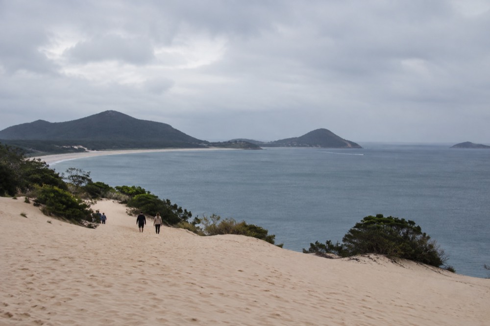 Tall pale sand dunes rising above calm bay water with distant green headlands
