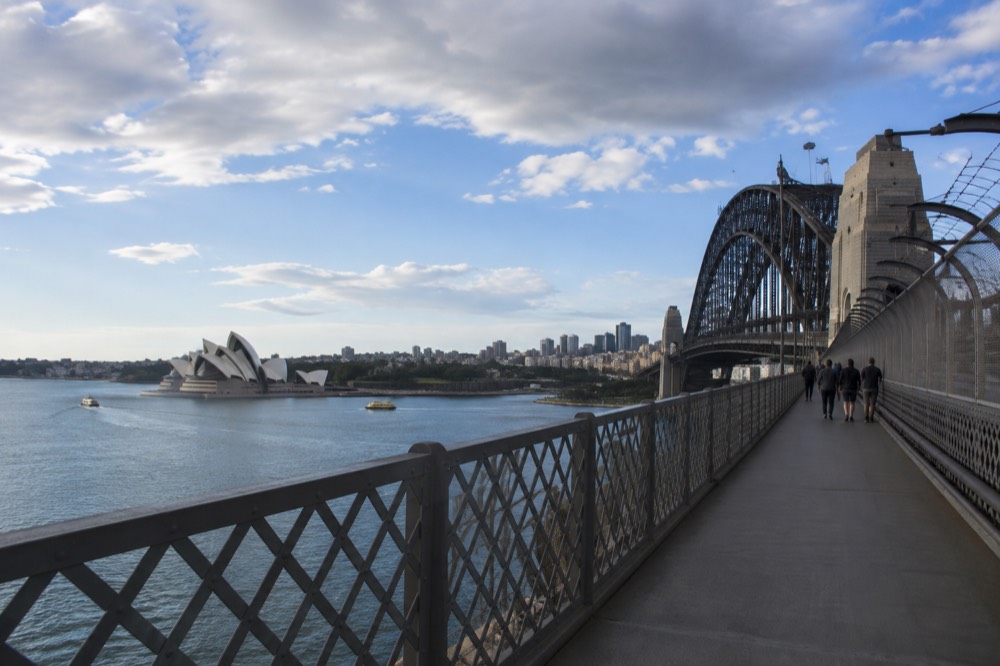 The eastern pedestrian walkway of the Sydney Harbour Bridge with the Opera House visible across the blue water