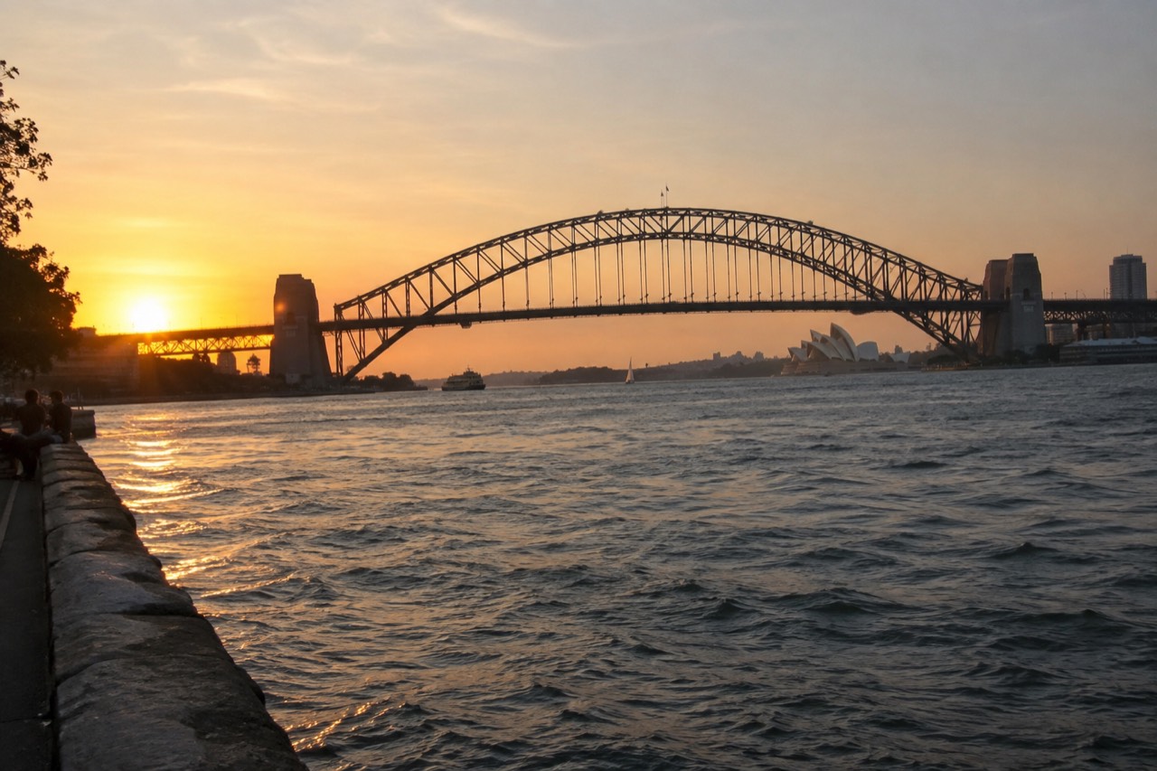 The Sydney Harbour Bridge seen from the harbour at dusk, with the Opera House lit across the water