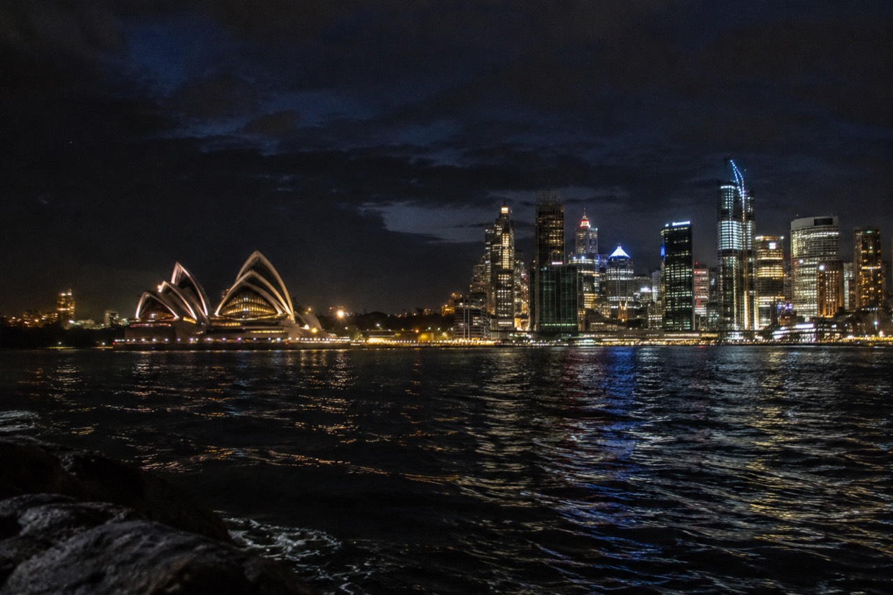 Sydney Harbour and city skyline lit up after dark, with the Opera House glowing beside the black water