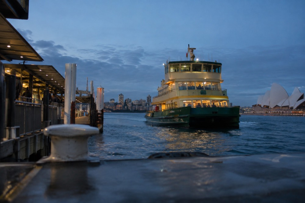 A green and yellow Sydney ferry approaching a wharf with harbour water in the foreground
