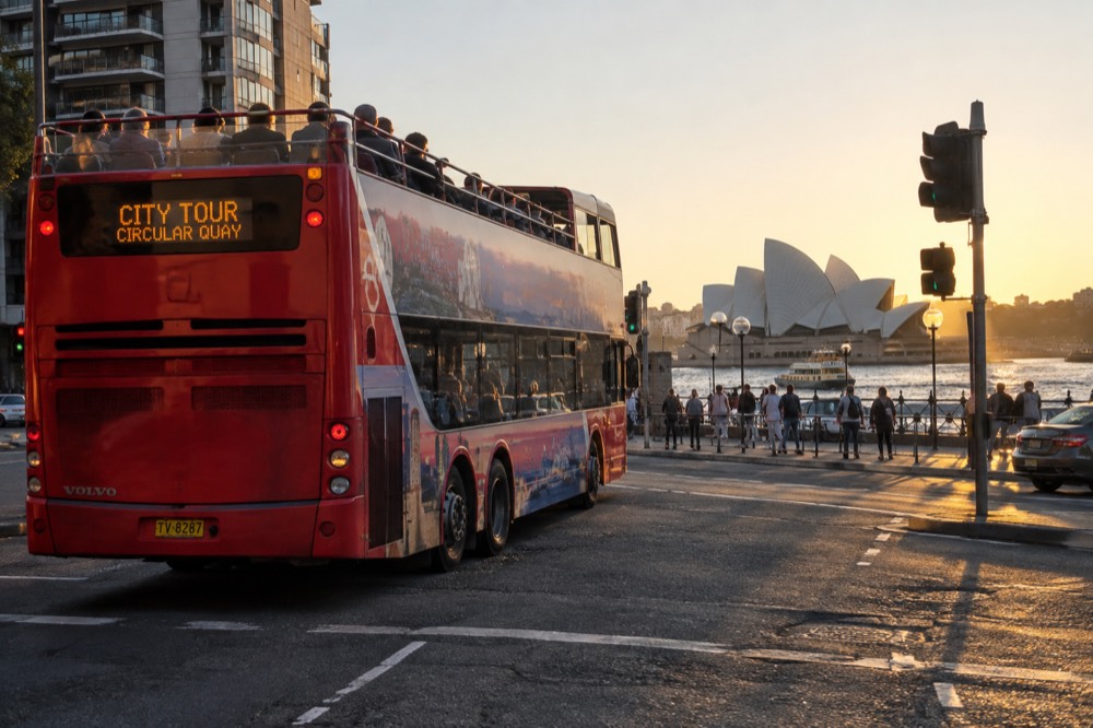 A double decker sightseeing bus crossing a city street with harbour buildings behind it