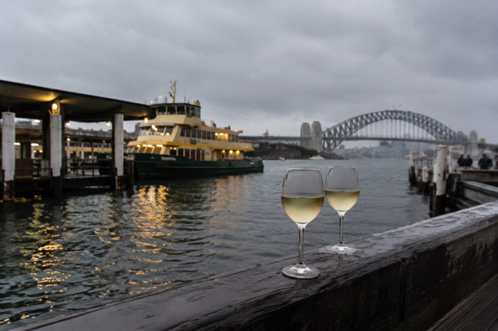 Two wine glasses on a timber jetty railing with ferry lights reflected on dark harbour water