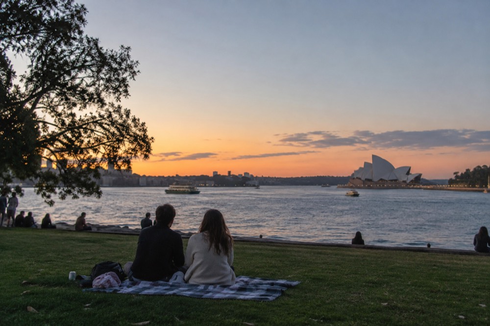 A picnic blanket on a green lawn overlooking Farm Cove with the Opera House in the distance