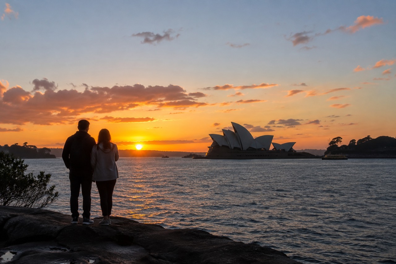 A couple standing at a harbourside lookout as the sun sets over Sydney Harbour and the Opera House silhouette