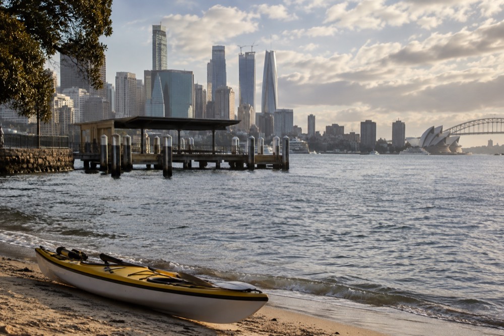 A kayak resting on a harbour beach with the city skyline in soft morning light