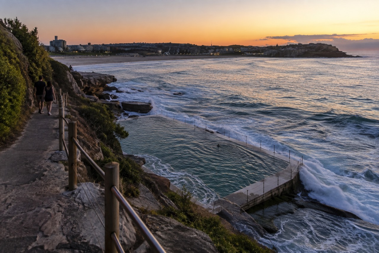 A sandstone cliff top path above an ocean pool with waves breaking on the headland