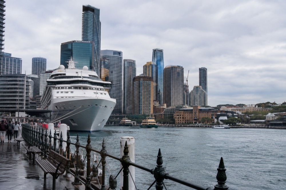 A white cruise ship moored at a Sydney harbour terminal with the city skyline behind it