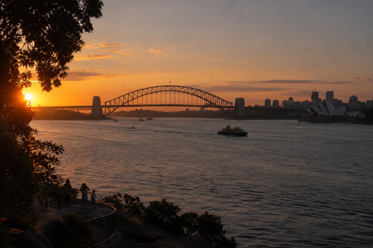 Sydney Harbour at dusk with the Opera House sails and Harbour Bridge silhouetted against an orange sky