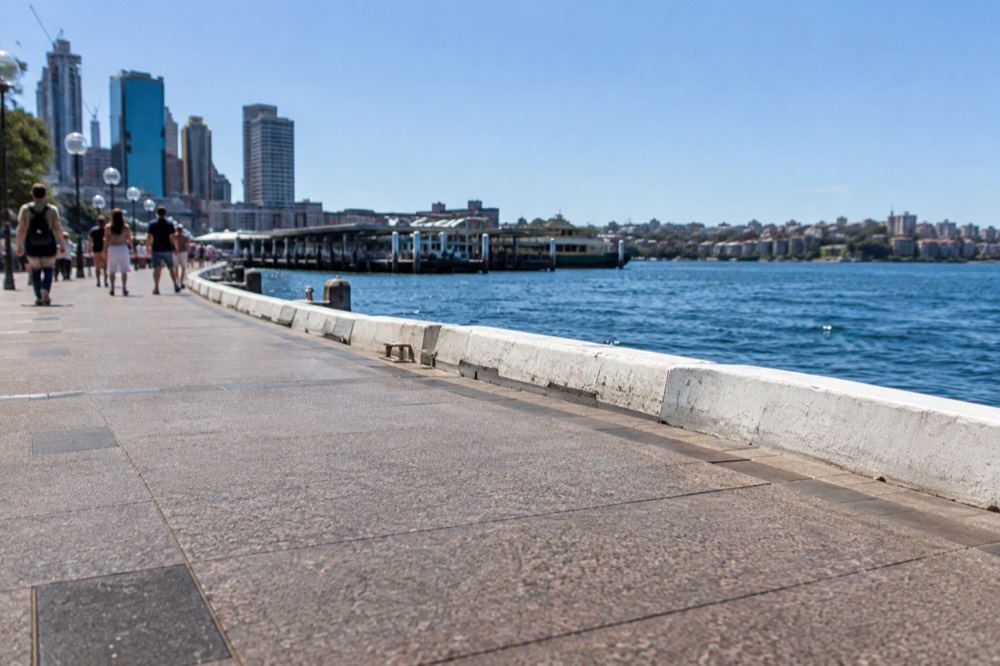 A wide harbourside path beside calm blue water with a ferry terminal in the distance