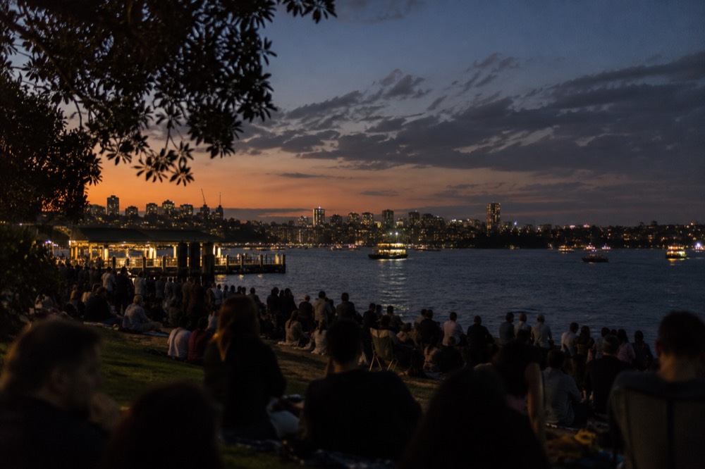 Crowds gathered on harbour foreshore grass at dusk waiting for the fireworks