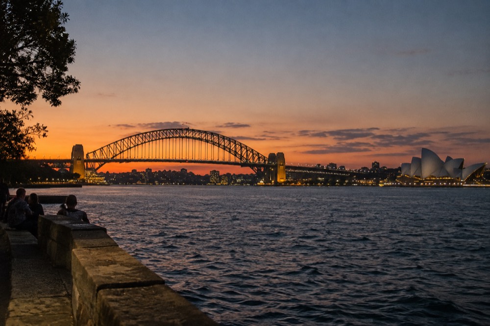 Sydney Harbour foreshore at dusk with the Opera House and harbour bridge lit against a calm evening sky