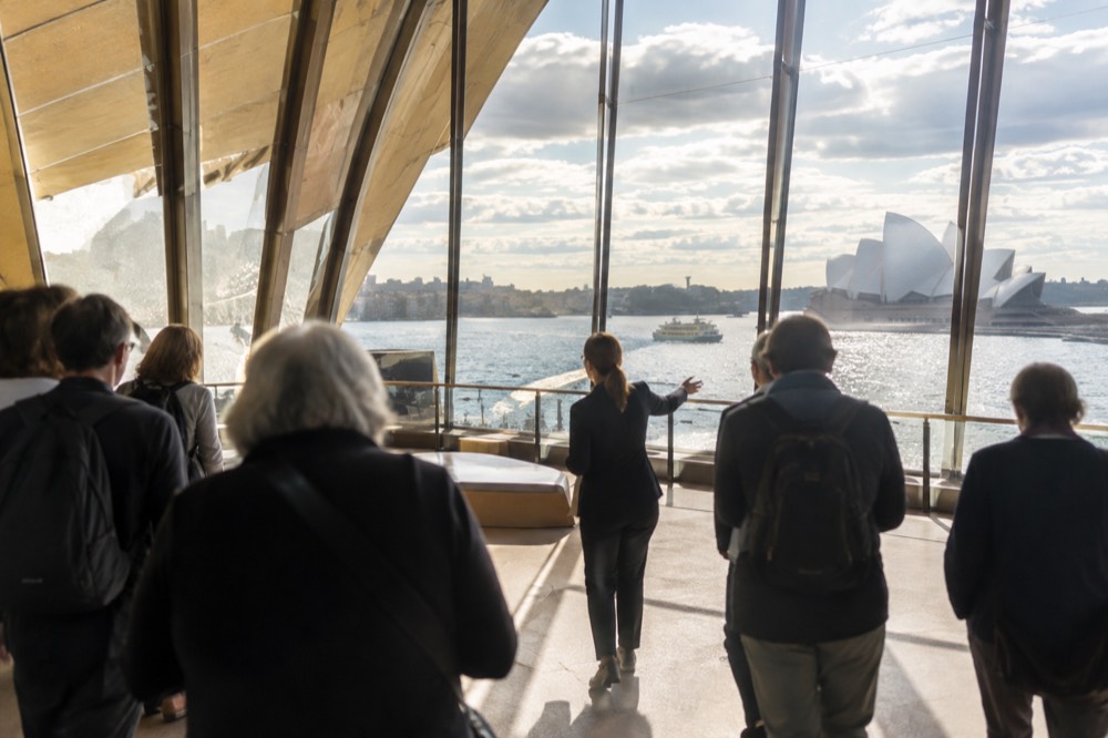 A guide leading a small group through a bright foyer of the Sydney Opera House with harbour views through tall windows