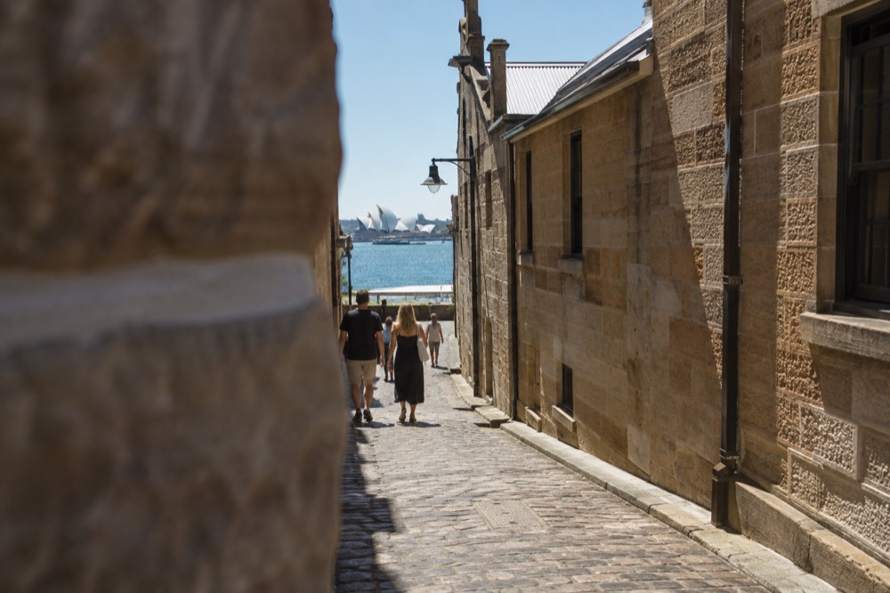 Cobblestone laneway between heritage sandstone walls leading toward the harbour