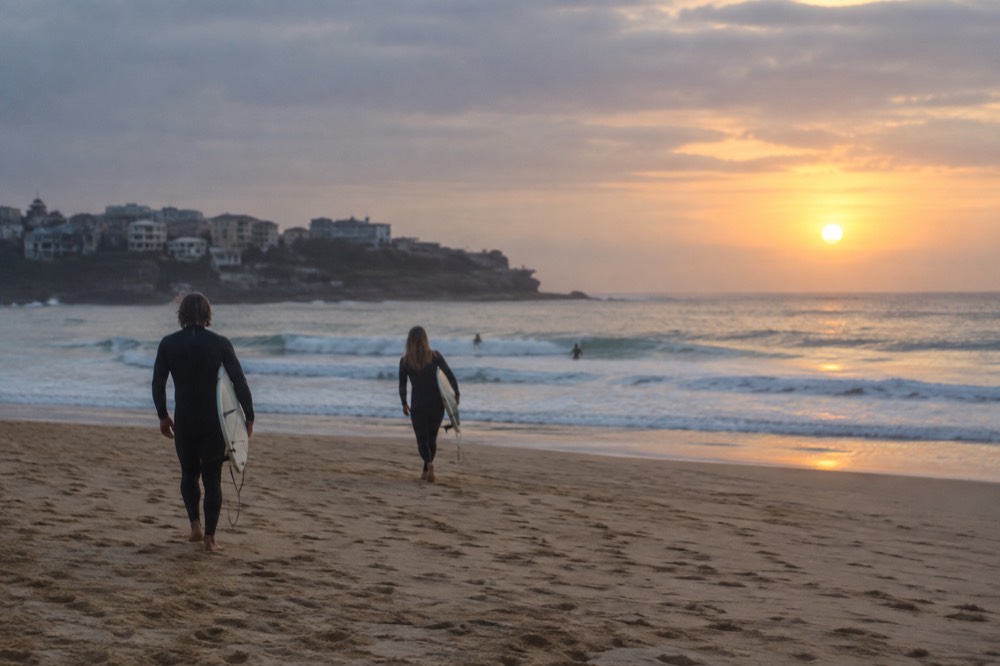 Surfers walking across golden sand toward small waves at sunrise