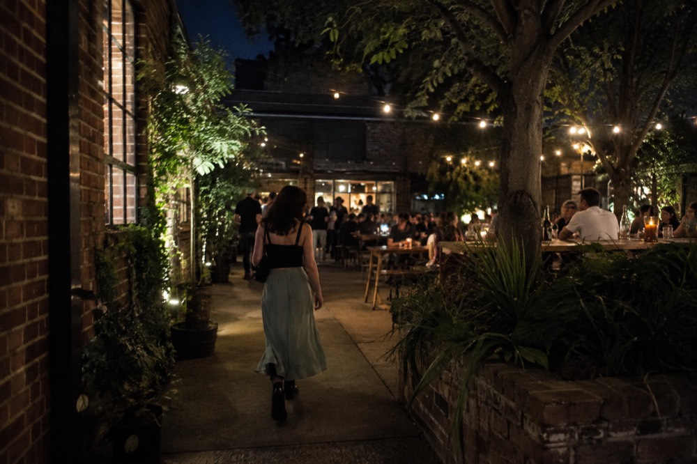 A courtyard with long shared tables and people holding drinks under string lights