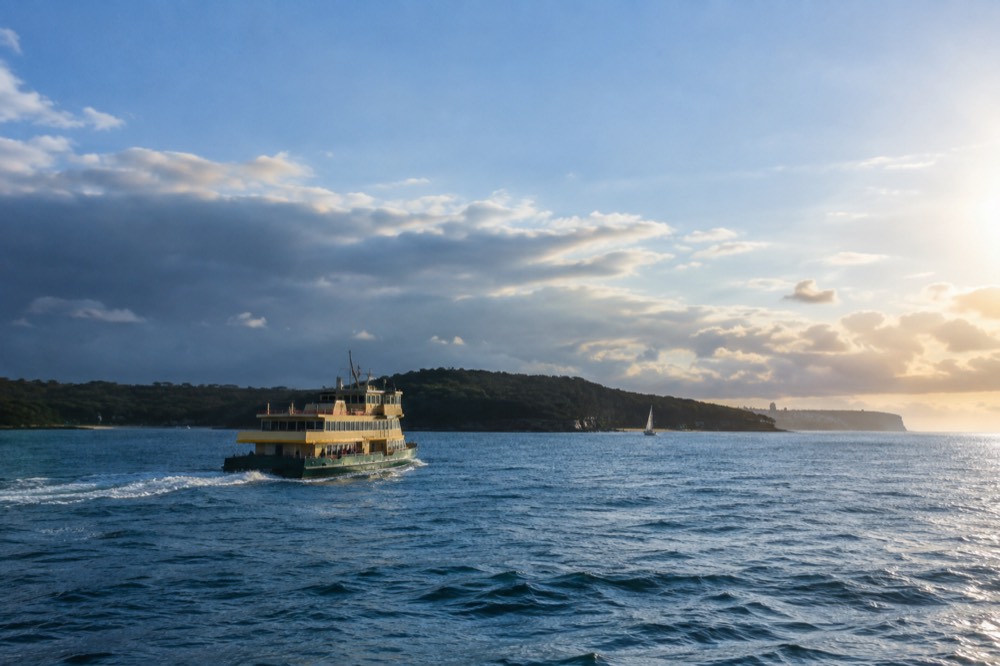 A green and cream ferry crossing open blue harbour water toward a treed headland