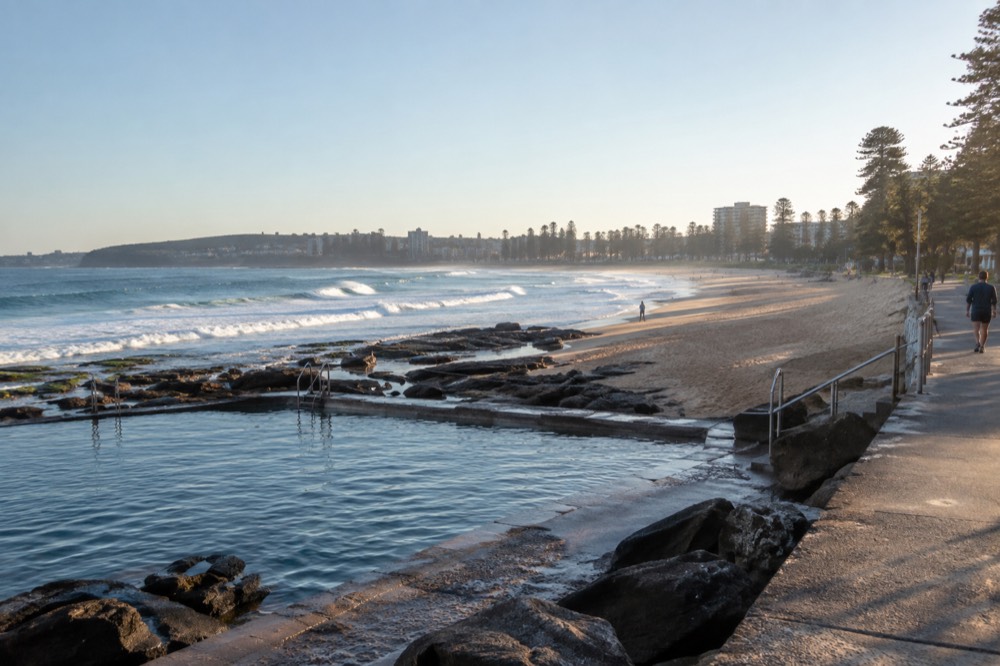 A long ocean beach with a rock pool at one end and a paved promenade behind the sand