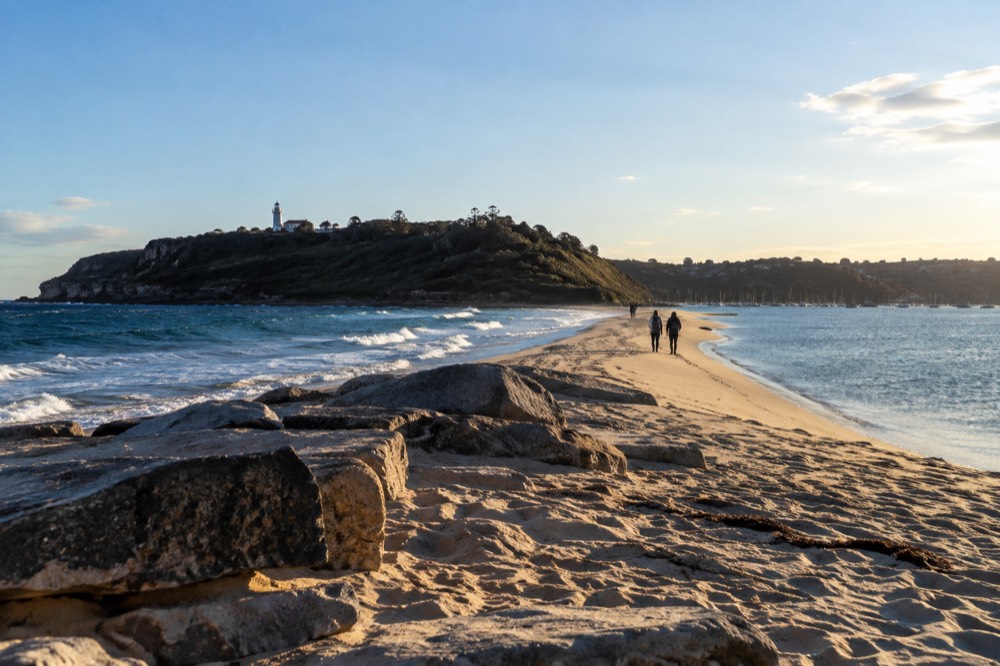 A narrow sand spit between dark ocean water and a calm blue inlet, with a lighthouse on the headland