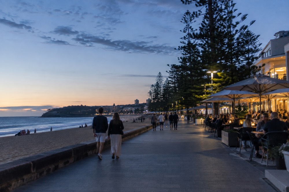 A beachside cafe strip with outdoor tables and people walking a paved promenade