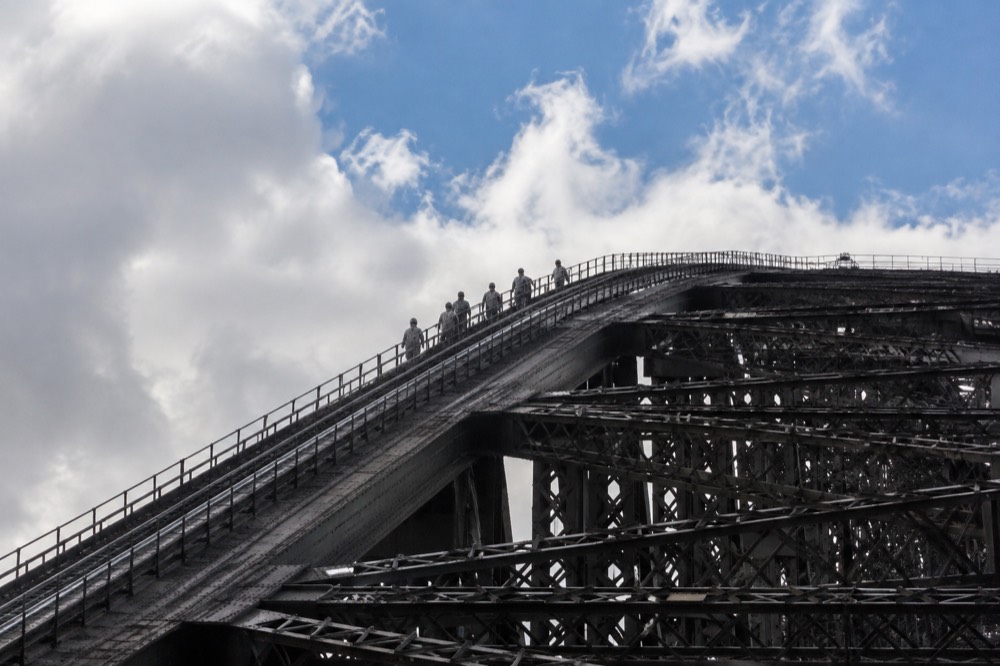 A group of climbers in grey jumpsuits walking along the upper arch of the bridge, clipped to the safety cable