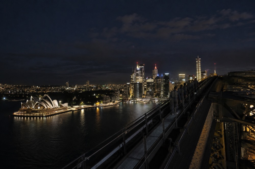 The Sydney skyline illuminated at night, seen from the upper arch, with the Opera House lit white against the dark water