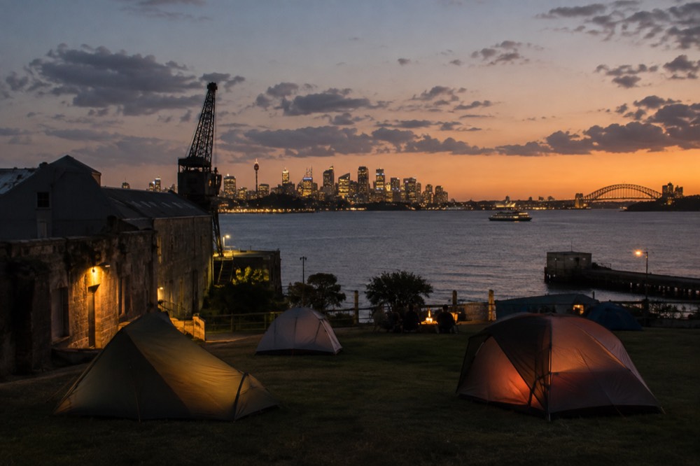Tents pitched on a grassy plateau on the island with the city skyline lit up across the water at dusk