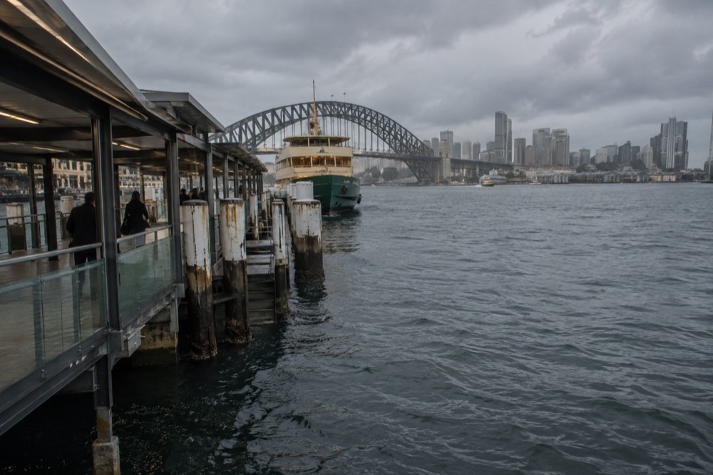 A ferry wharf on a harbour with a city skyline and bridge behind it
