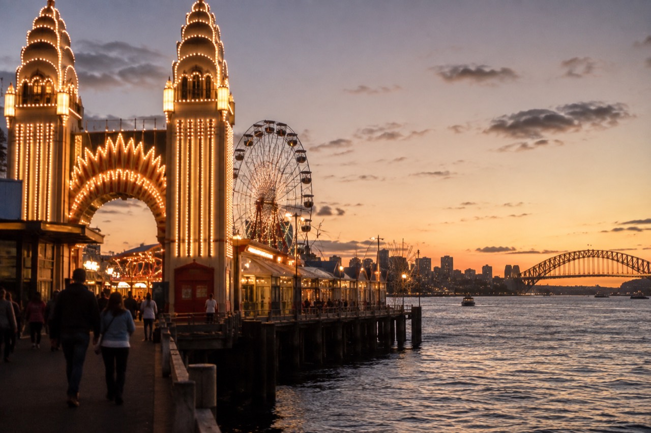 The illuminated entrance face of a vintage funfair beside a harbour at dusk
