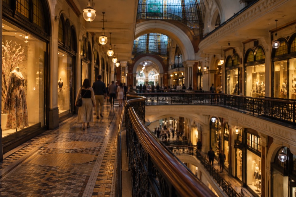Shoppers strolling past illuminated shopfronts beneath an ornate ceiling in a heritage arcade