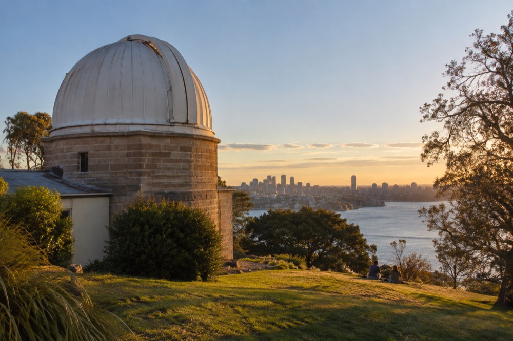A white heritage observatory dome against a blue sky above a green city hilltop
