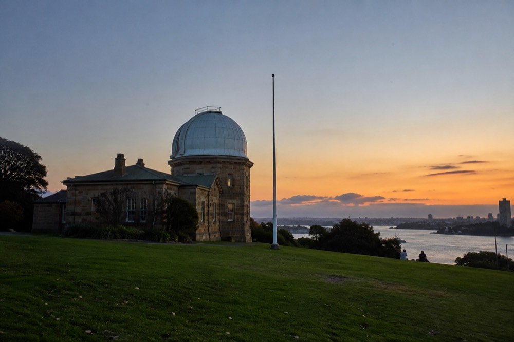 A sandstone heritage observatory building with a white dome beside a flagpole on a green hill