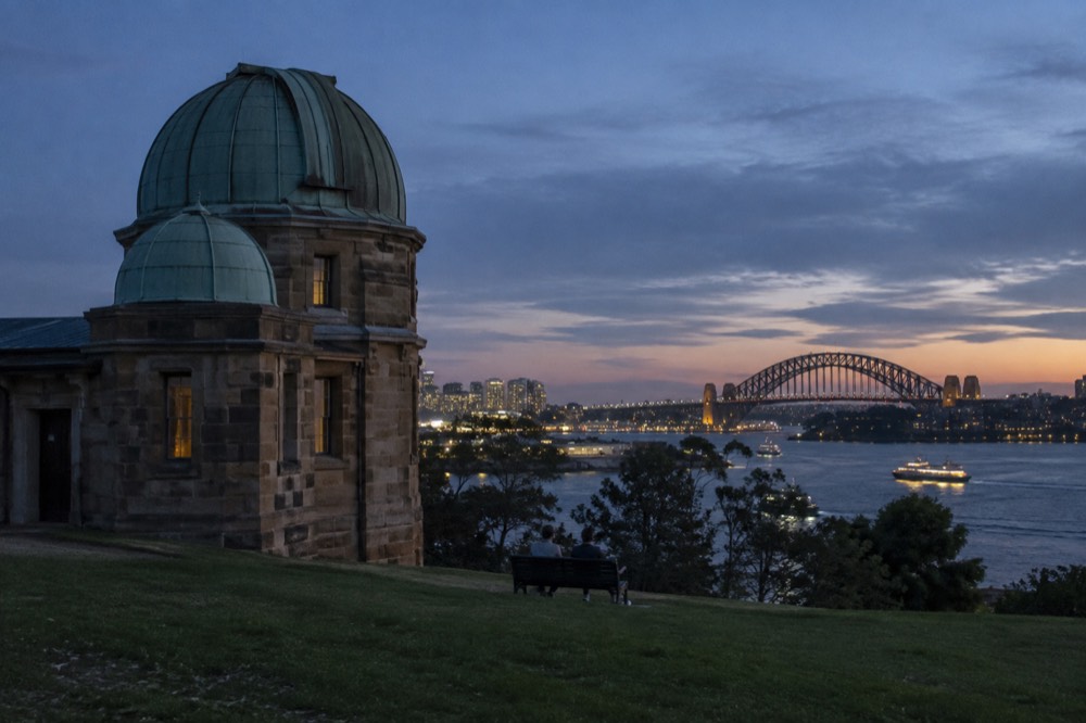 An evening sky over a harbour city with a lit-up steel arch bridge and ferries crossing dark water