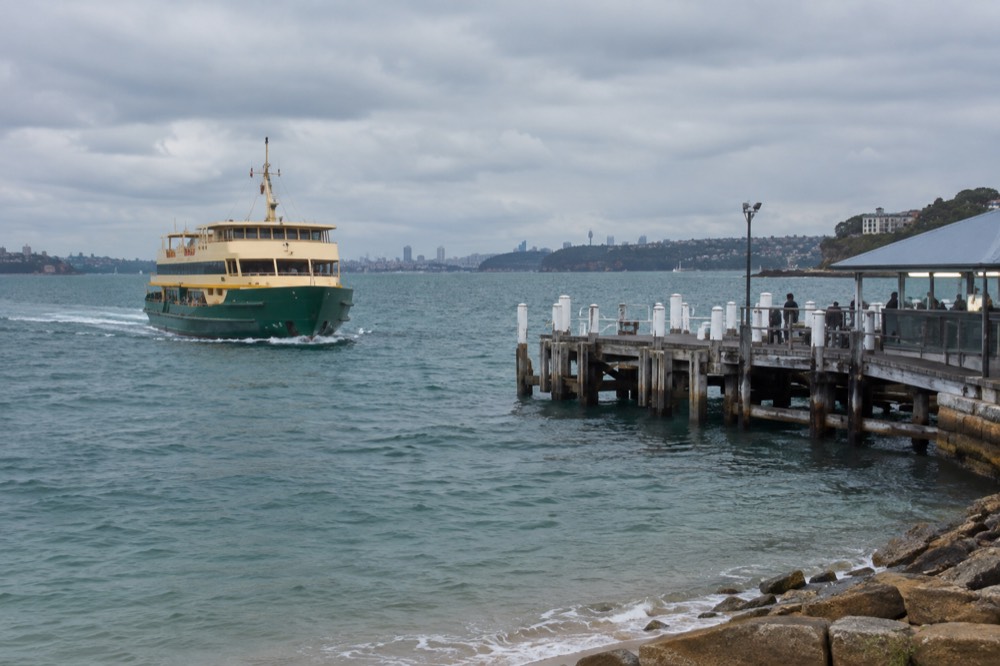 Green and cream ferry approaching a timber wharf on sparkling harbour water