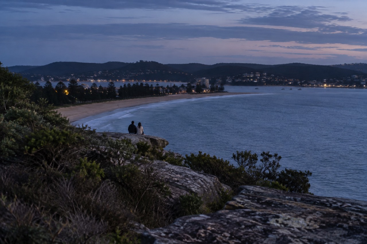 Sandstone headland above a long curved beach with calm blue water and a distant town
