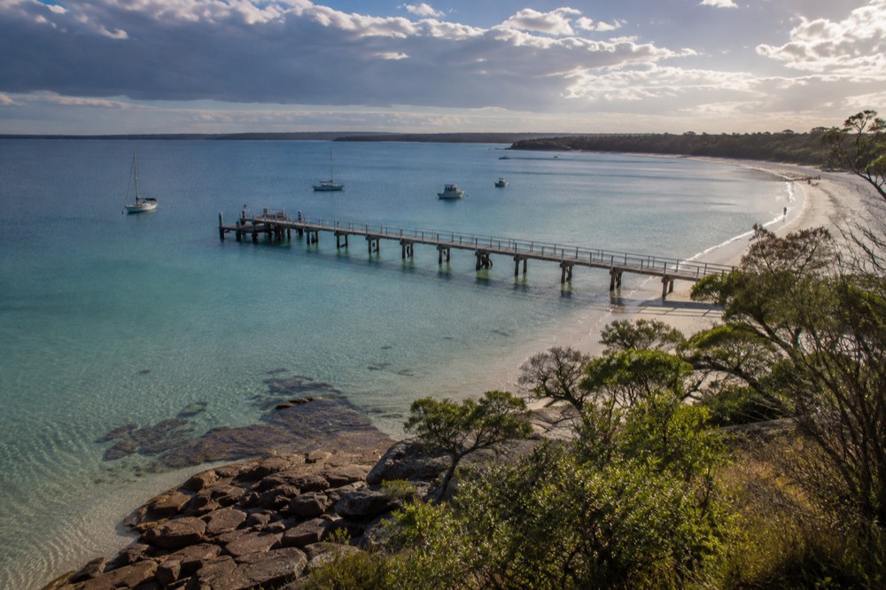 A calm blue bay with white sand and a wooden jetty reaching into clear water