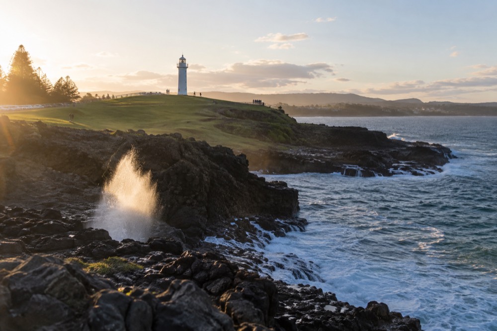 A white lighthouse on a green headland above a blue ocean bay