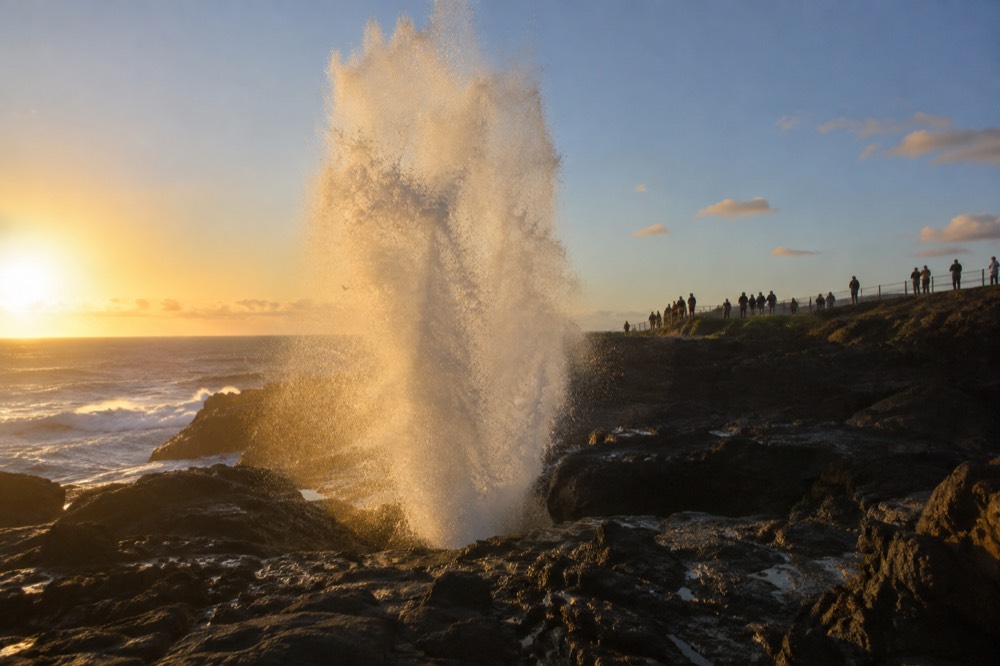 Ocean spray erupting vertically from a rock channel on a headland while onlookers stand back
