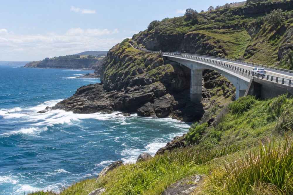 A curved road bridge clinging to green cliffs above a blue ocean