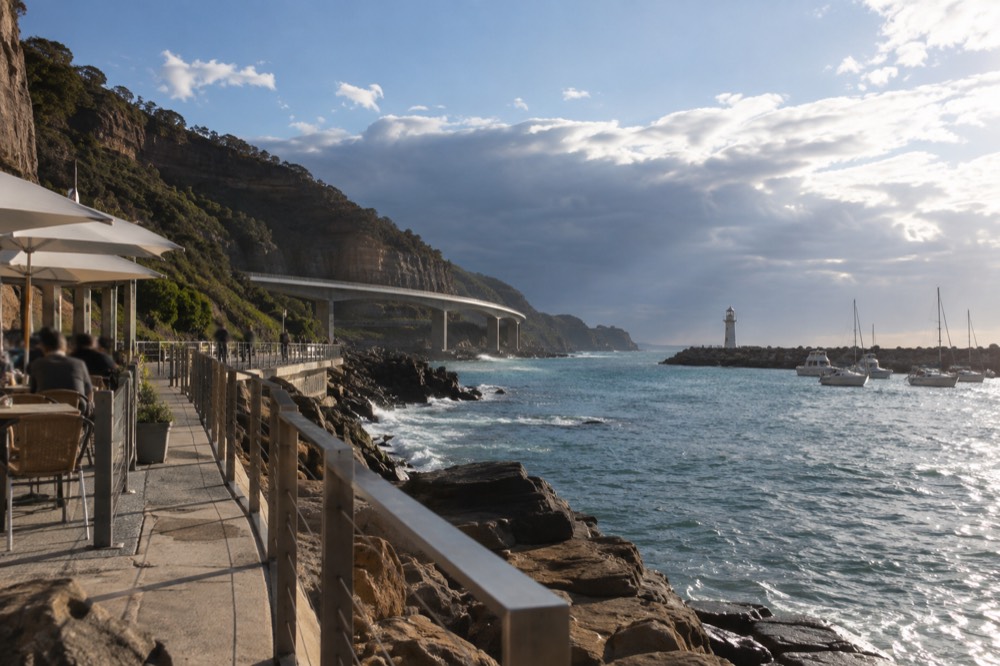 A waterfront cafe strip beside a boat harbour with a lighthouse in the distance