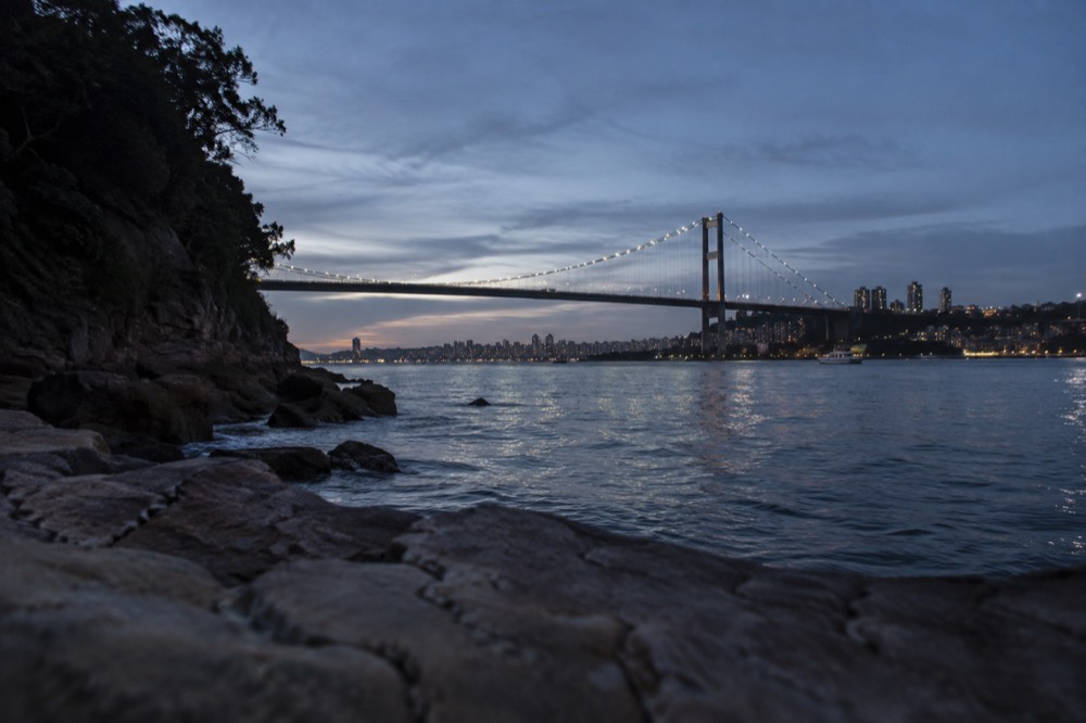 A suspension bridge across a harbour at dusk with city lights beginning to show