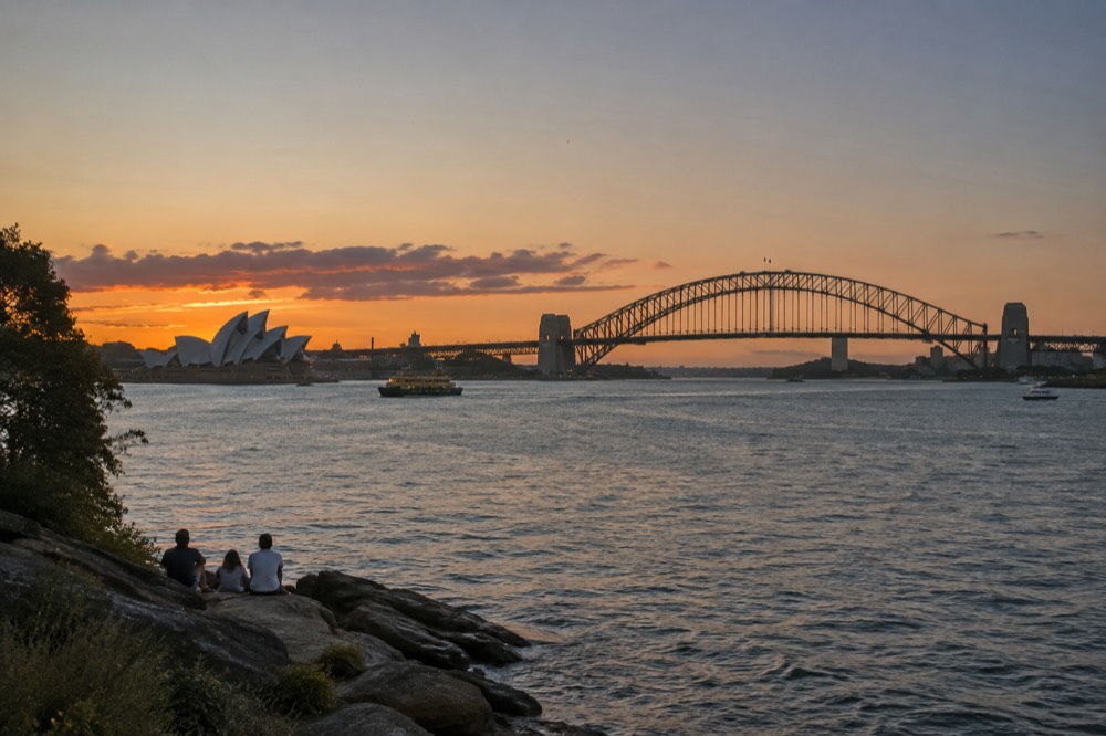 Sydney Opera House and Harbour Bridge seen across the water from a harbour headland at golden hour