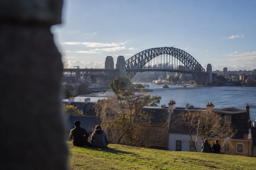 A grassy hilltop above old sandstone rooftops with the Harbour Bridge rising behind